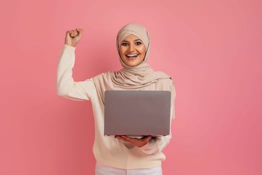 Joyful muslim woman in hijab holding laptop and shaking her fist