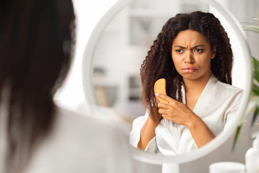 Upset Black Lady Combing Hair With Bamboo Brush Near Mirror At Home