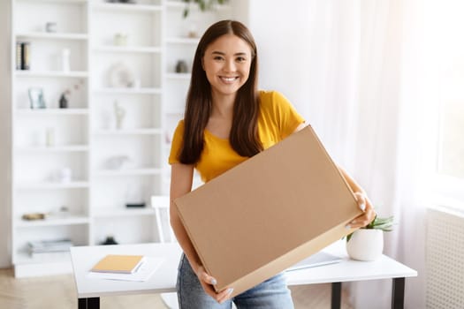Happy Asian woman holding large cardboard parcel in bright home office