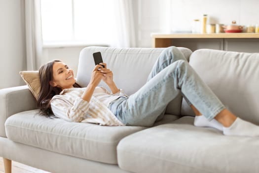 Joyful young woman with smartphone in hands lying back on soft sofa