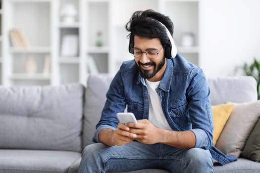 Relaxed indian man listening music on smartphone with wireless headphones at home