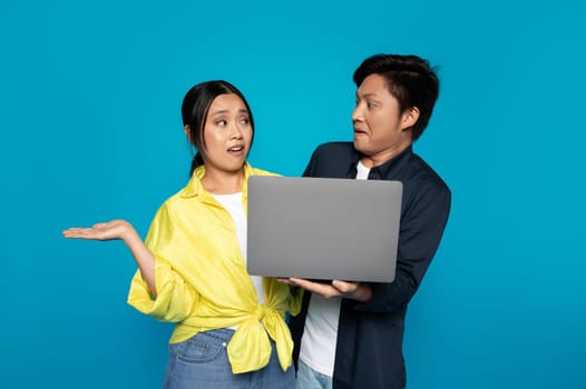 Confused Asian woman gesturing with an open hand while looking at a laptop held by a focused man