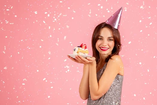 Delighted woman in a sparkling dress and party hat, holding a piece of birthday cake