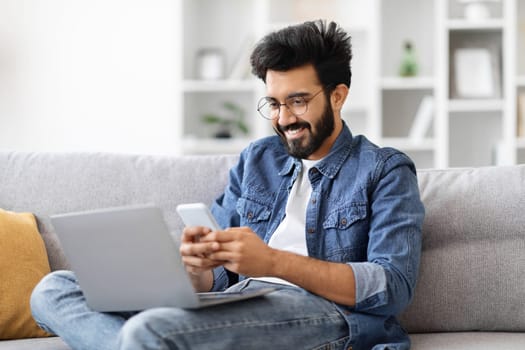 Engrossed indian man in eyeglasses multitasking with laptop and smartphone at home