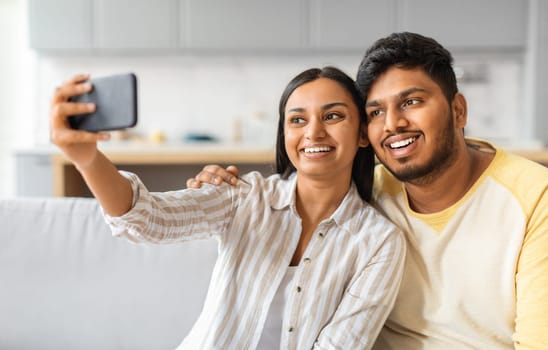 Cheerful young indian couple sitting on sofa and taking selfie on smartphone