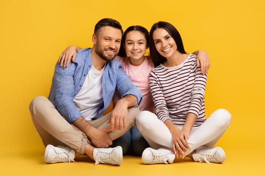 Portrait Of Happy Young Family Of Three With Teen Daughter. Cheerful European Parents And Their Cute Teenager Female Kid Sitting On Floor Over Yellow Studio Background And Smiling At Camera