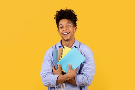 Content black male student hugging notebooks on yellow backdrop