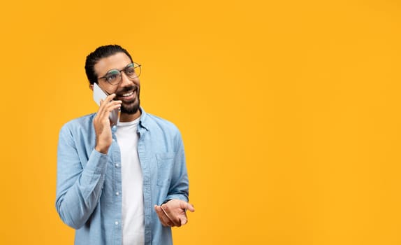 A man with a beard and glasses cheerfully talks on the phone, gesturing with his free hand
