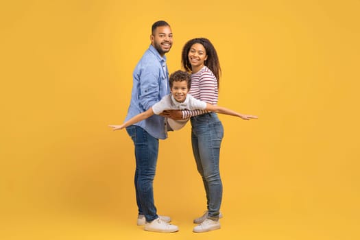 Family Fun. Cheerful Black Mom And Dad Playing With Their Preteen Son, Bonding Together Over Yellow Studio Background, Happy African American Boy Pretending Flying, Spreading Hands, Imitating Plane