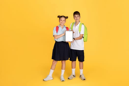 A smiling boy and girl in school uniforms, with backpacks, stand side by side holding a tablet