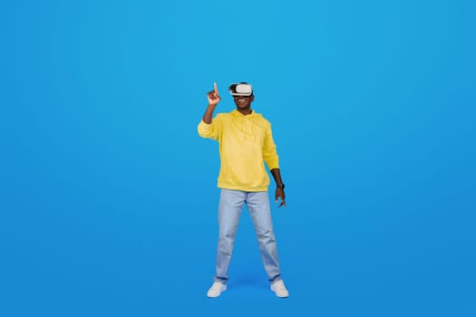 A Black man in a yellow hoodie points upwards while wearing virtual reality goggles