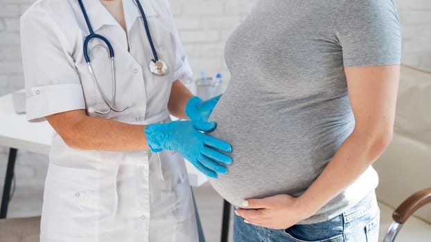 Pregnant woman visiting a doctor. Elderly Caucasian female gynecologist holds hands on the tummy of a pregnant patient.