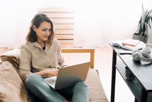 Cheerful millennial woman typing on a laptop, attending a virtual meeting while seated on a sofa with a gadget. Joyful millennial woman typing on a laptop, attending a virtual meeting seated on a sofa