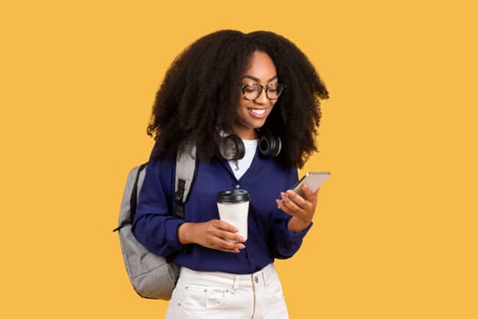Excited african american female student using smartphone, wearing backpack and headphones, holding coffee to go