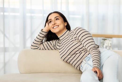 Pensive glad young arab woman lies on sofa in light living room