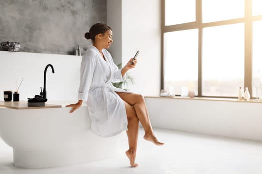 Black woman in white bathrobe laughing while texting on her smartphone