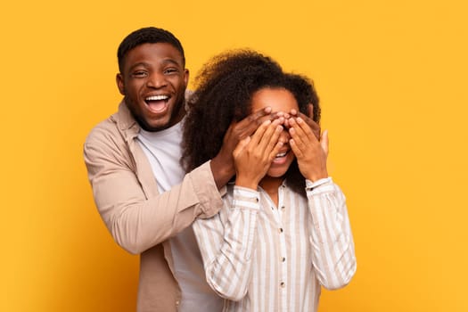 Black man covering woman's eyes for surprise on yellow background