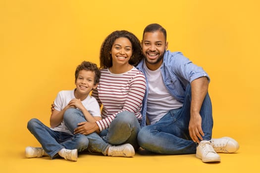 Black family with preteen son sitting on floor and smiling towards camera, happy young african american parents and their male child embracing and bonding, posing isolated on yellow background
