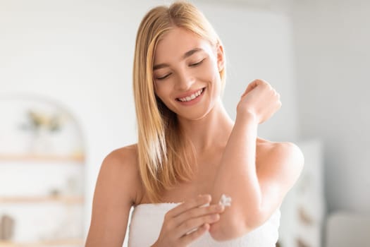 Attractive blonde woman applies moisturizer on elbow in bathroom