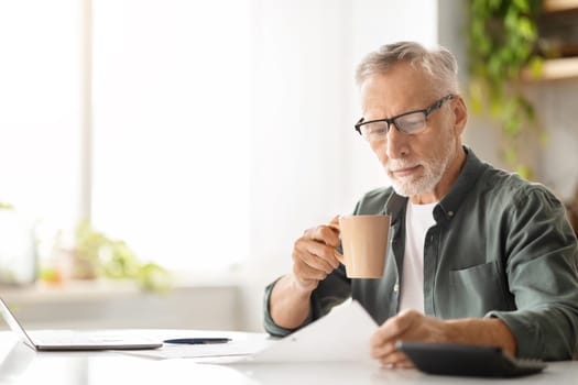 Concentrated elderly male in glasses drinking coffee while reviewing documents at home