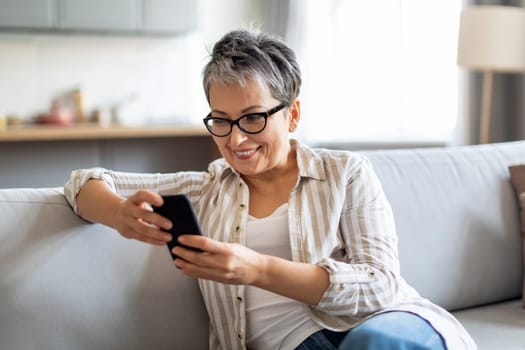Smiling Senior Woman Using Smartphone While Relaxing On Couch At Home