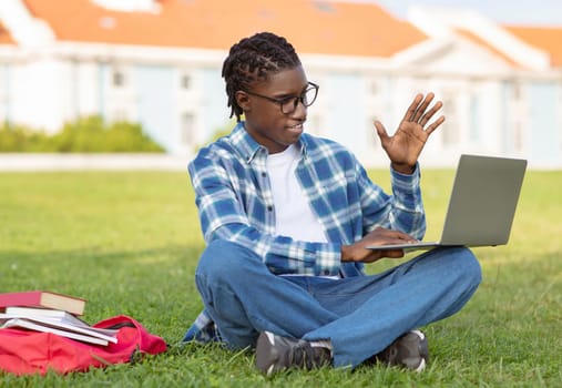 Happy black student guy studying on laptop waving hello outdoor