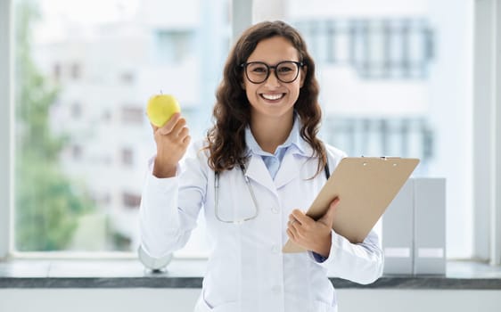 Cheerful young woman doctor holding apple and clipboard