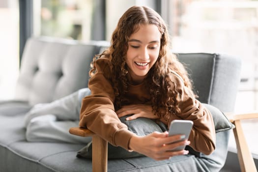Smiling caucasian youngster girl using her mobile phone for communication, relaxing on sofa at home interior, texting and browsing web on smartphone gadget. Modern teen lifestyle