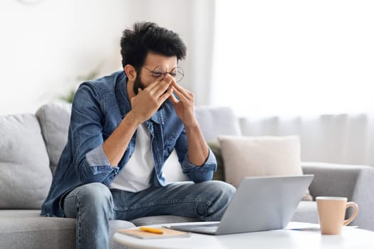 Tired indian man rubbing eyes, suffering fatigue while working on laptop
