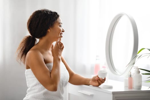 Black beautiful woman applying moisturising cream on face while looking in mirror