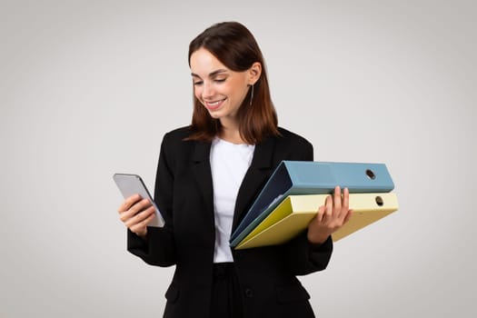 Happy busy young European woman in suit with smartphone chatting in app, folders