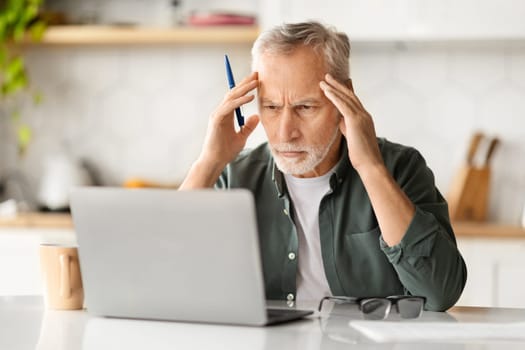 Portrait of stressed senior man using laptop in kitchen interior