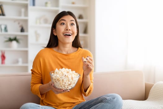 Amazed young chinese woman watching new TV show, eating popcorn