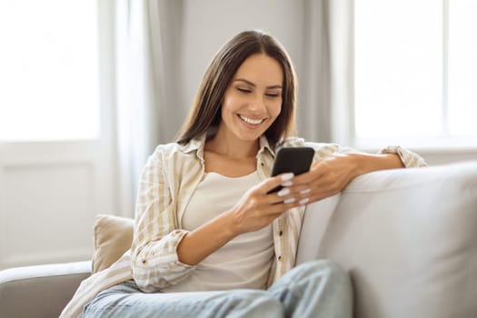 Relaxed young woman smiling and browsing her smartphone while reclining on couch