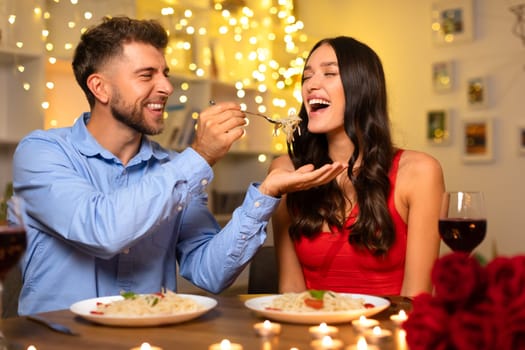 Joyful man feeding pasta to laughing woman during romantic dinner