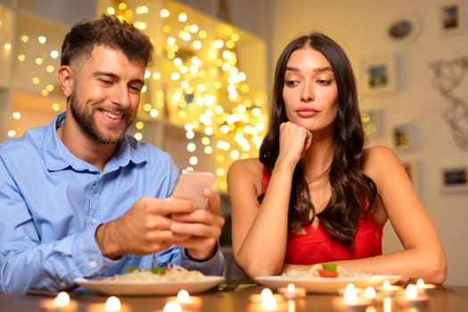 Man on phone smiling, woman bored at candlelit dinner