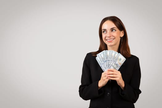 Smiling young European lady in suit hold many money cash and look at copy space