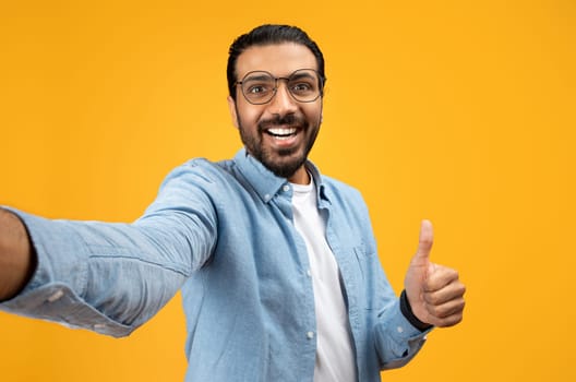 An exuberant man with a beard and glasses takes a selfie, giving a thumbs up with a beaming smile