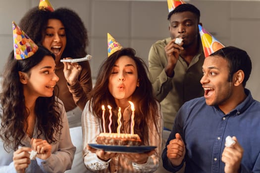 Multiracial friends looking at bday woman blowing out candles indoor