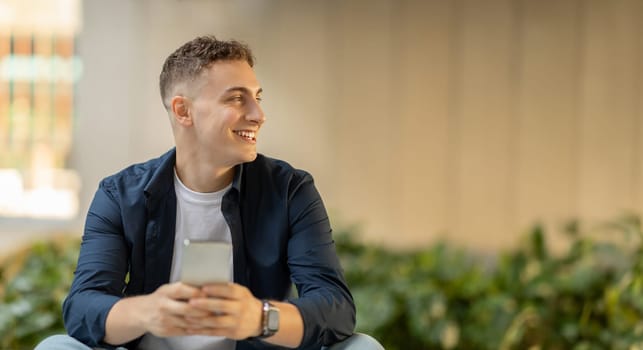 A cheerful young man looking away with a smile, holding a smartphone