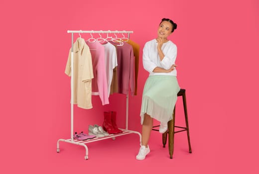 Fashion Idea. Pensive Smiling Asian Woman Sitting Near Clothing Rack