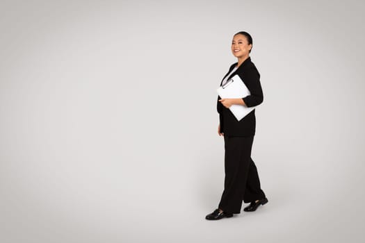 Smiling confident young asian woman in suit with clipboard walk