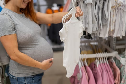 Caucasian pregnant woman chooses baby clothes in a store. Faceless expectant mother in the 3rd trimester.
