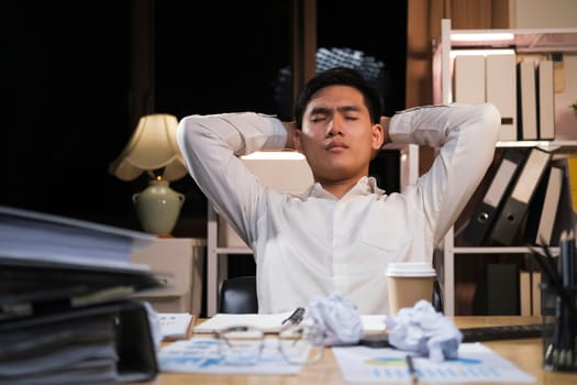 Asian man working late sitting on desk in office overtime at night. Business man feeling tired and stress for overload job.