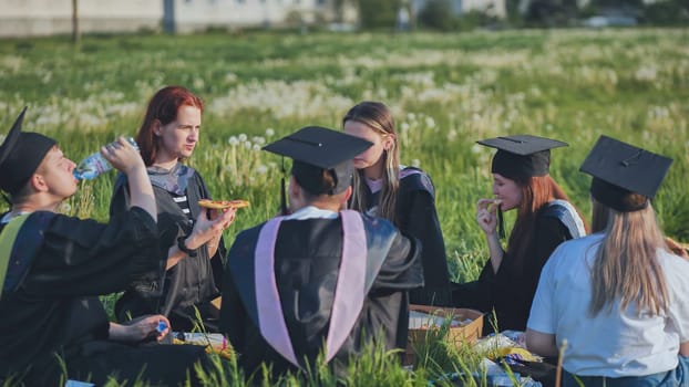 Graduates in black suits eating pizza in a city meadow.