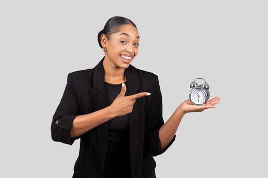 Smiling surprised millennial black businesswoman in suit points finger at alarm clock she holds
