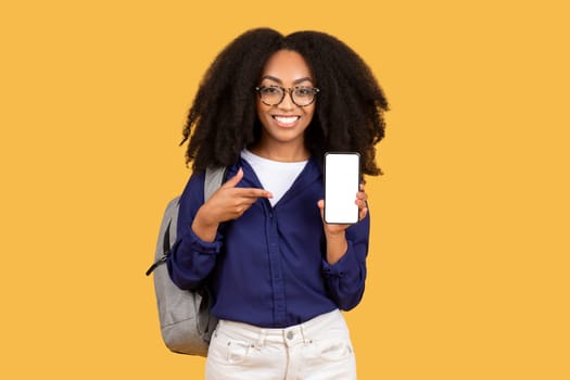 Happy young black lady with glasses pointing to smartphone with blank screen, perfect for app promotion, against yellow background, mockup