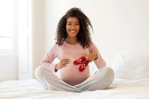 Maternity Concept. Black pregnant female embracing belly and holding small baby socks
