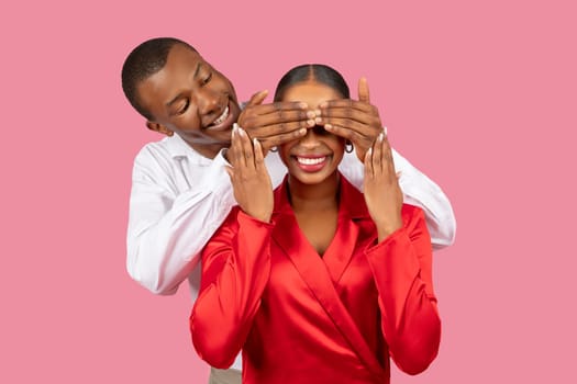 Black man covering woman's eyes for surprise on pink background