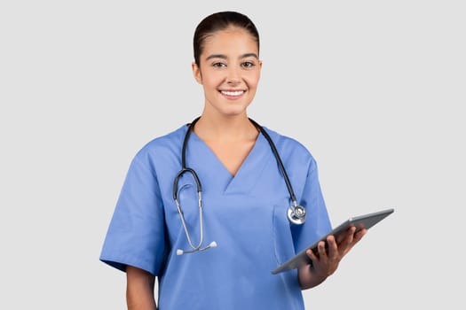A poised European millennial doctor in blue scrubs holds a tablet, her expression professional